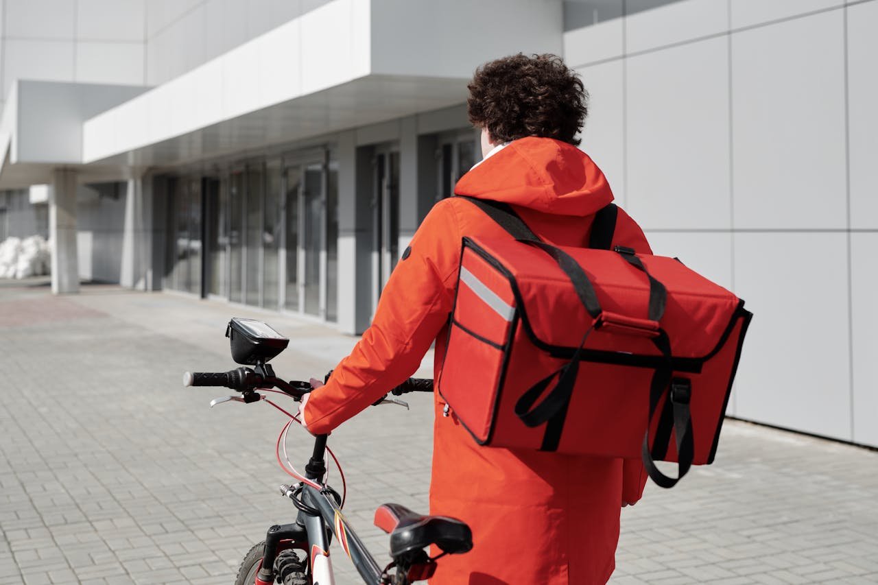 A food delivery rider in a red jacket walking a bike with a thermal bag outdoors.