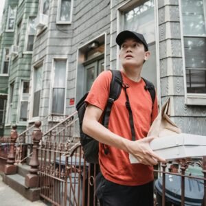 A delivery worker in a red shirt and cap holding packages outside a residential building.